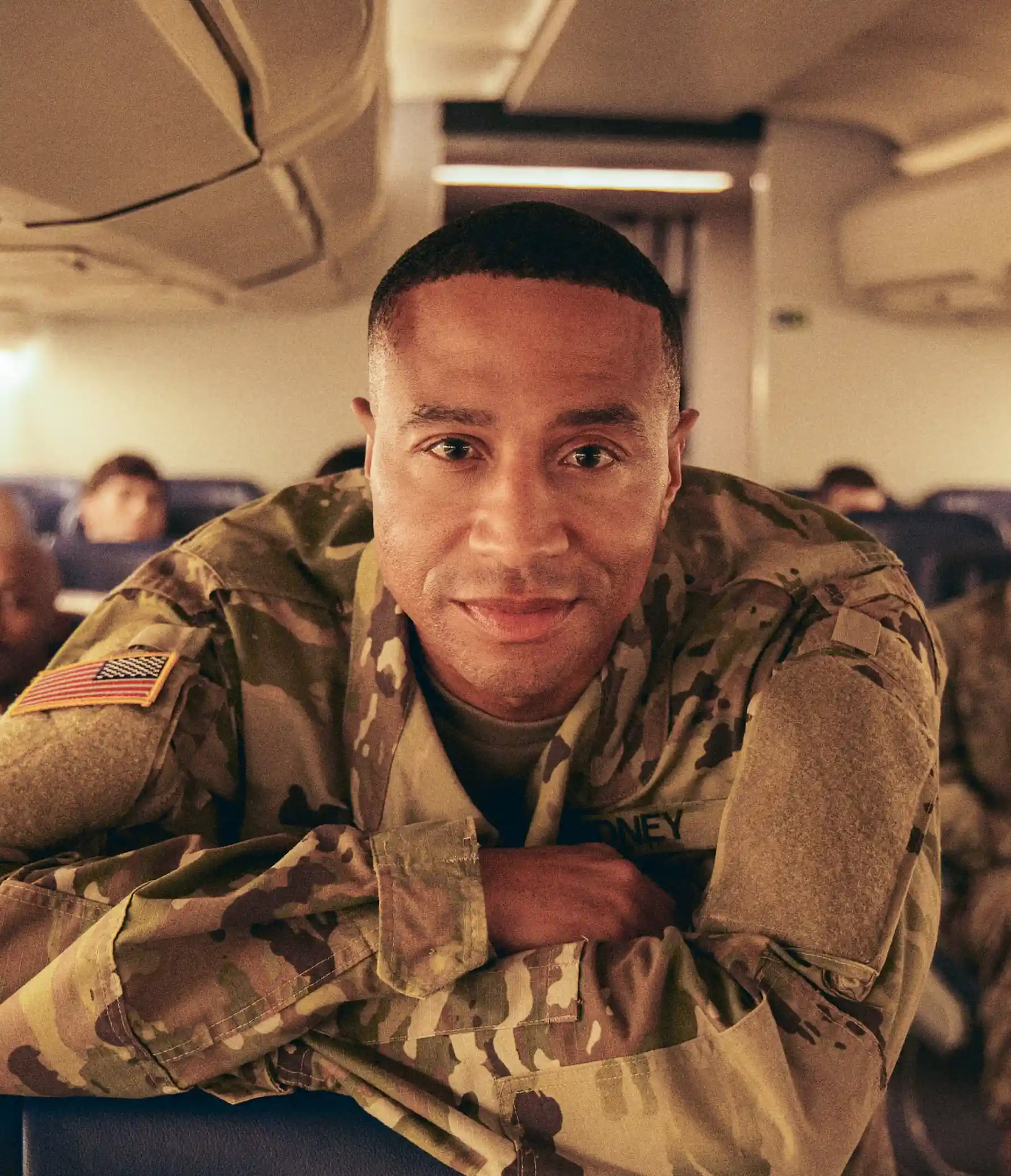 A Black soldier in a military uniform leans over the top of an airline seat on a flight with other soldiers.