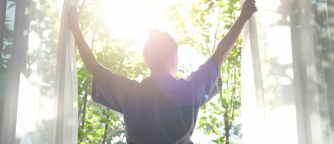 A woman holding curtains apart stands in the sunlight admiring nature through a window.