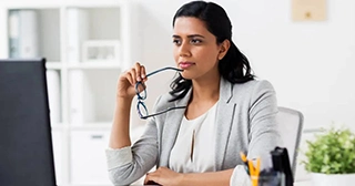 A woman with dark hair wearing a gray blazer holds her eyeglasses while contemplating a computer screen.