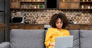 A young Black woman in a yellow shirt sits on a gray couch looking at a laptop.