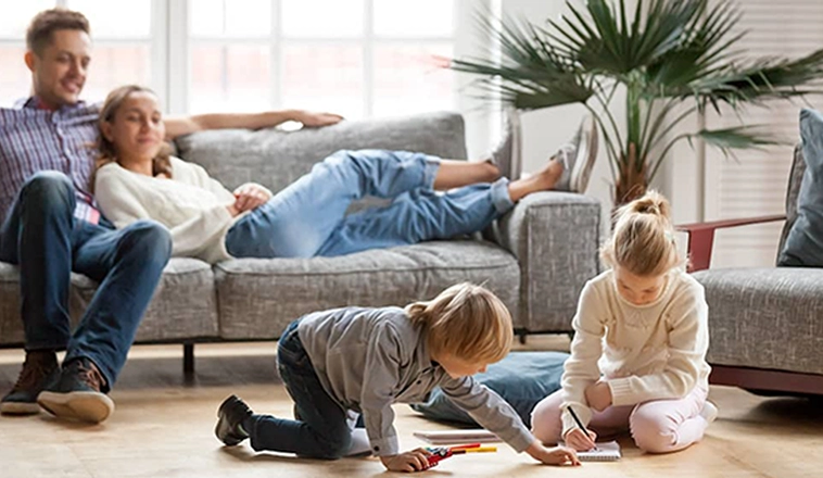 Two young parents in jeans lounge on a gray couch while watching their two children drawing together on the floor.