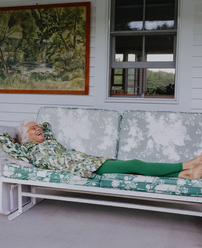 A woman with white hair relaxes on a porch swing in front of a white house that she purchased with Rocket Mortgage.