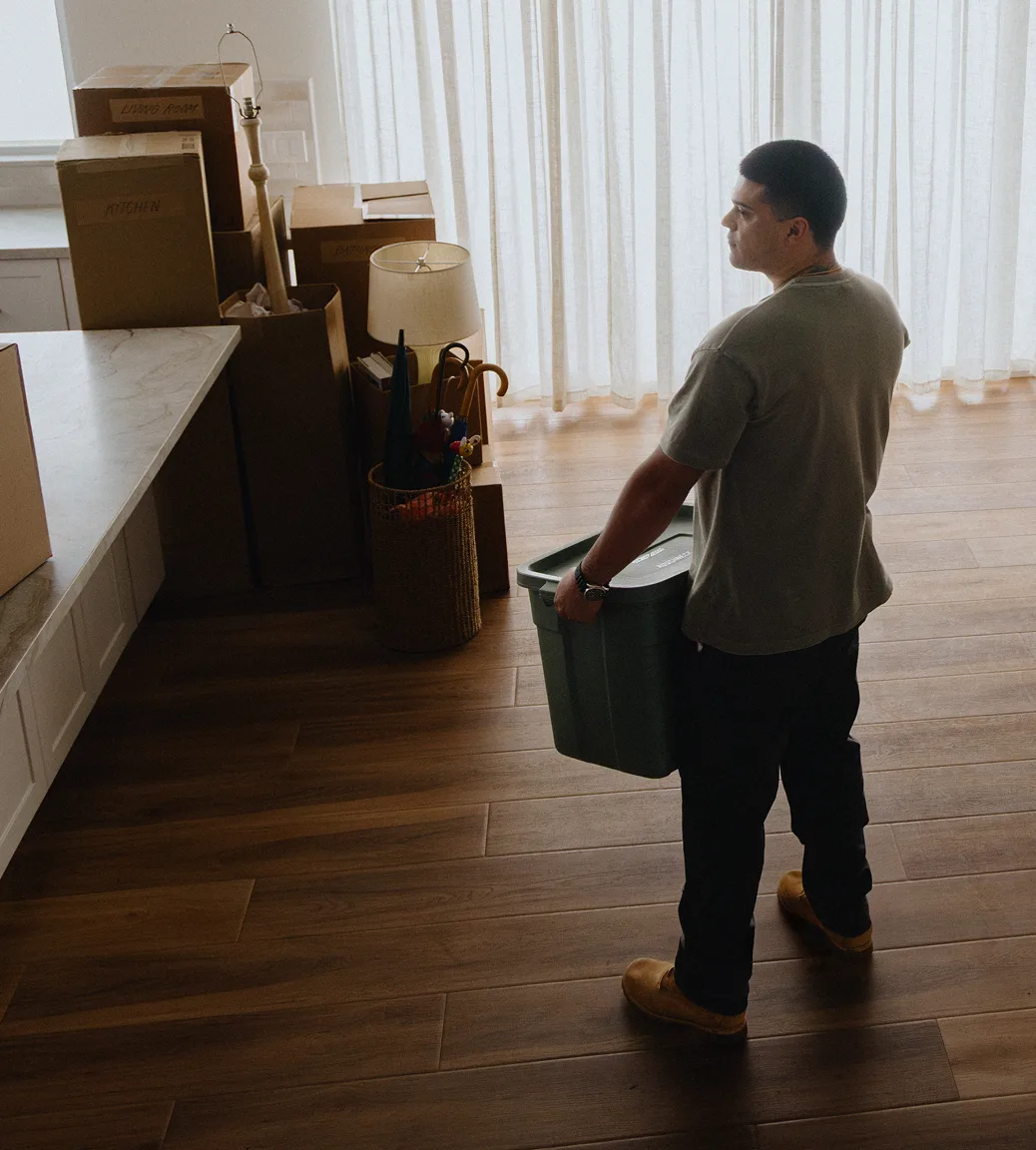 A man stands inside of his new home, holding a bin and surrounded by moving boxes.