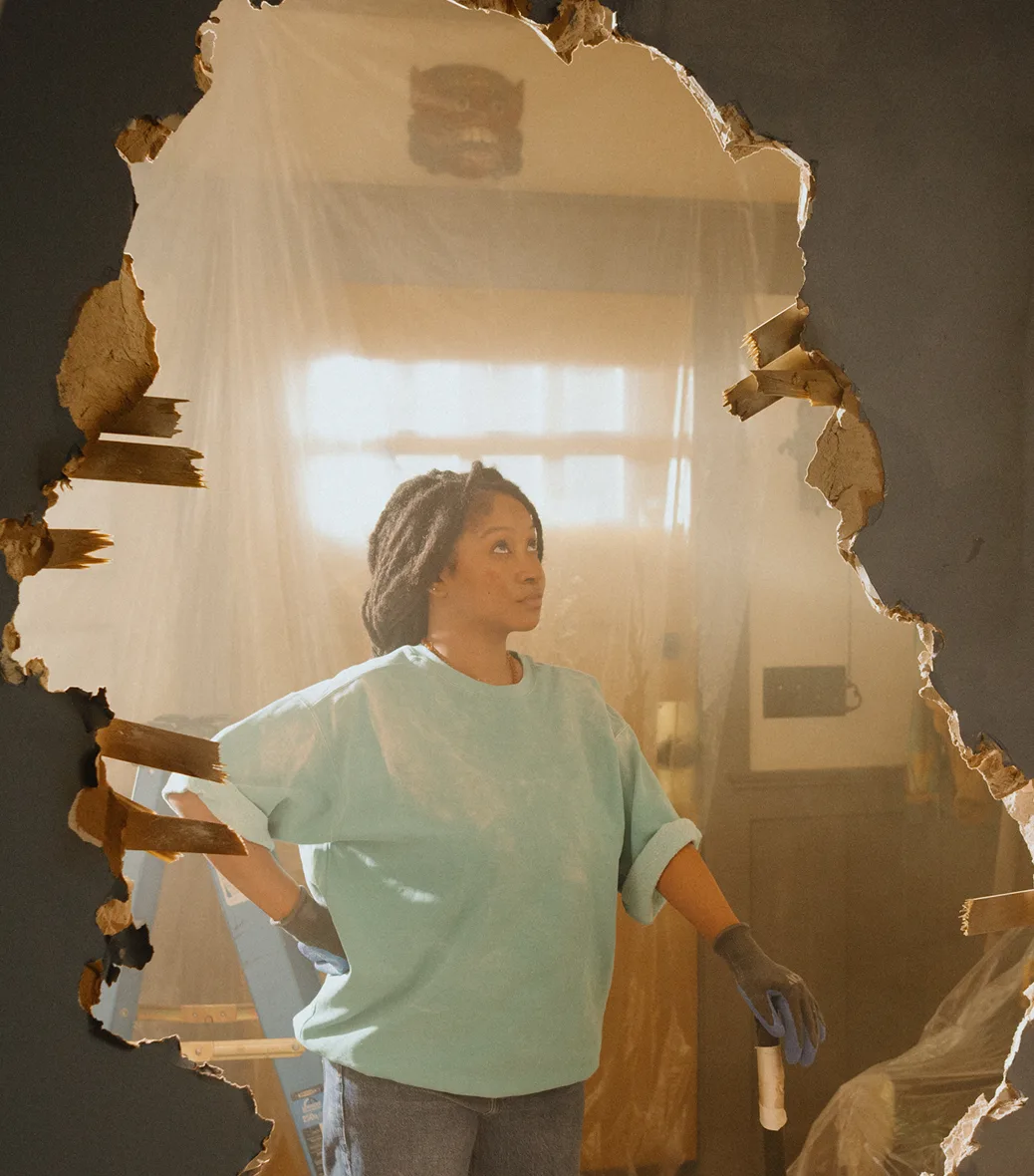 A woman in a blue sweatshirt stands in front of a large hole in a wall during a home renovation.