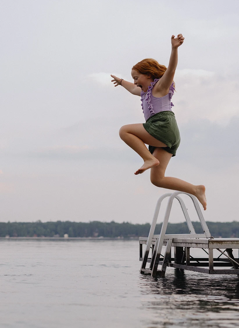 A young girl jumps off a dock into a lake, arms raised in excitement.