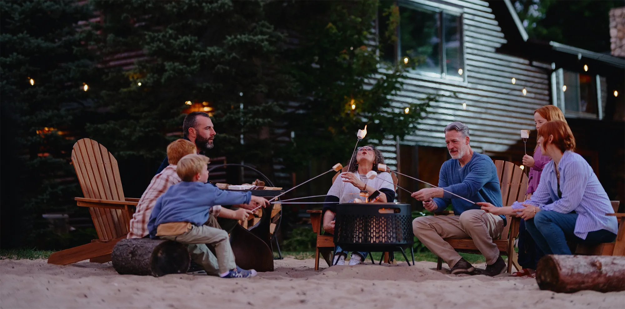 A multi-generational family sit around a fire pit outside their lake house, roasting marshmallows together in the evening.