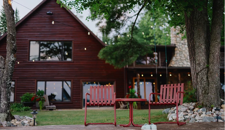 Two red chairs and a table on a dock overlook a lake, with a red cabin and large tree in the background.