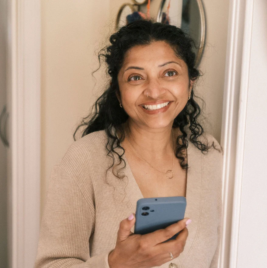 A woman in her home looks forward with her phone is in her hand.