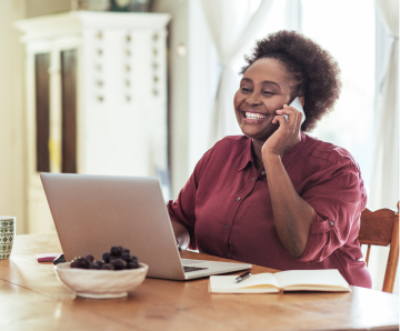 African American woman smiling while on her phone and sitting at her computer.