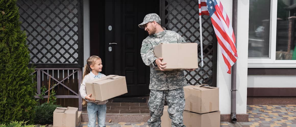 RHB Assets From IGX: Father in military uniform and daughter holding cardboard boxes near a house with an American flag.