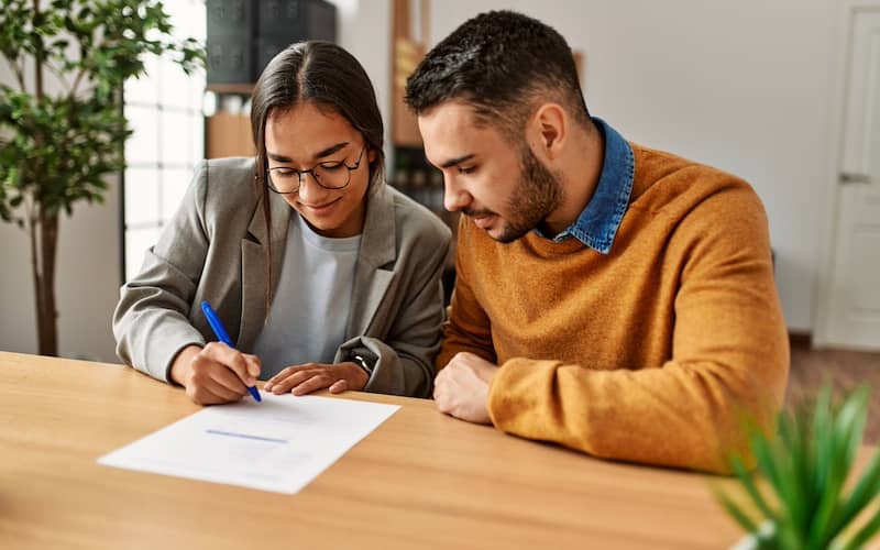 Woman in glasses and blazer and man in orange sweater sign document.