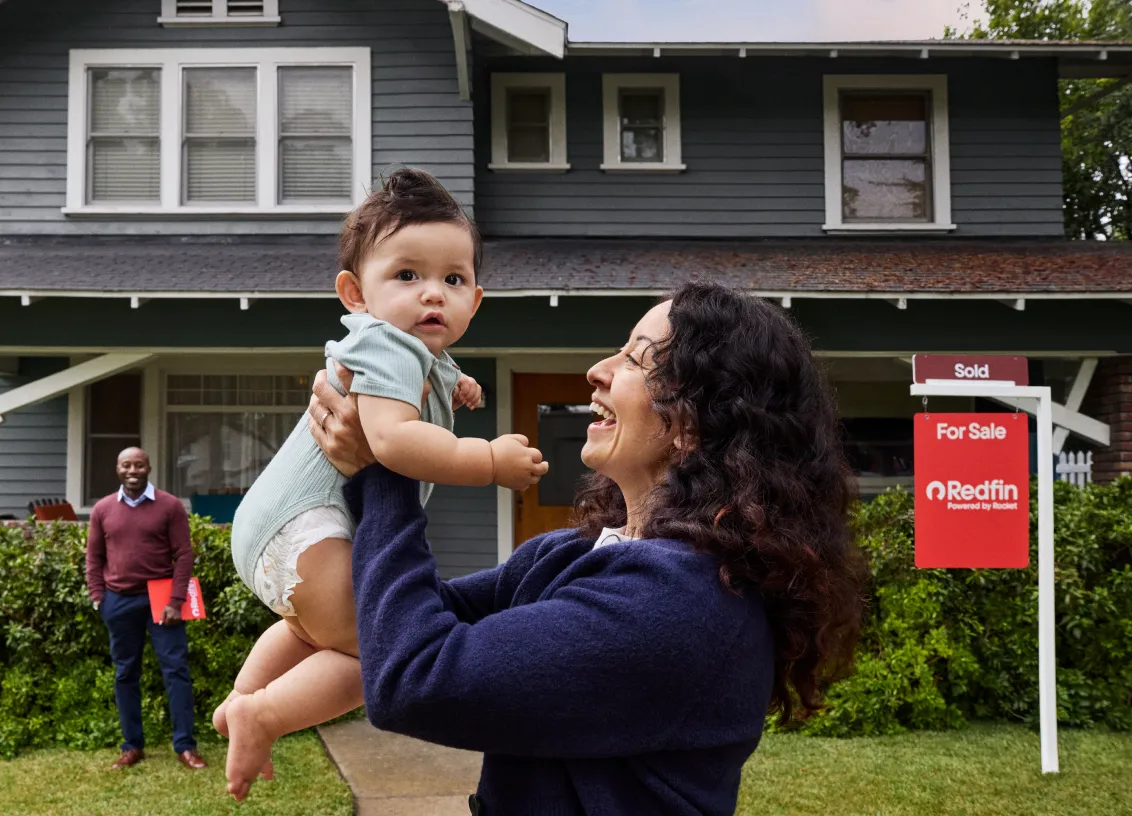 A woman lifting a baby in front of a green two-story house with a Redfin “Sold” sign in the yard while the realtor stands behind them.