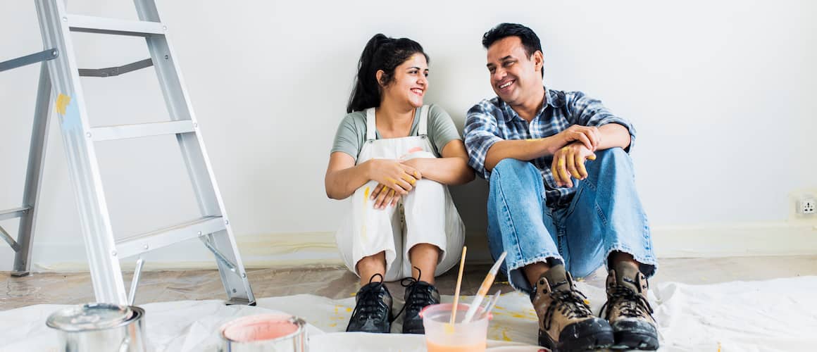 Two adults sit on the floor smiling at each other with opened paint cans and a ladder near them.