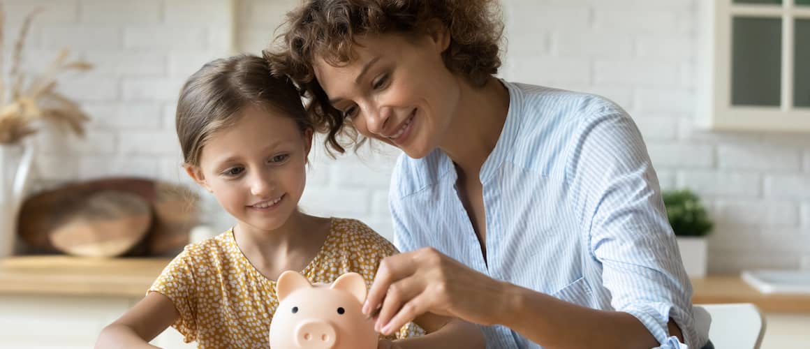 Mother and daughter adding coins to a piggy bank in their kitchen, smiling.