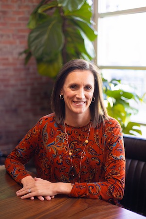 Headshot of a woman smiling, sitting at a wooden table.