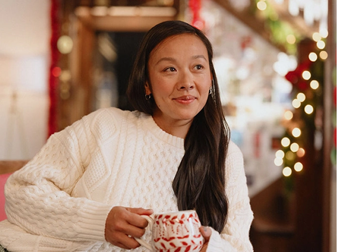 A woman in a white sweater holding a red-and-white patterned mug in a holiday-themed setting with lights and decorations.