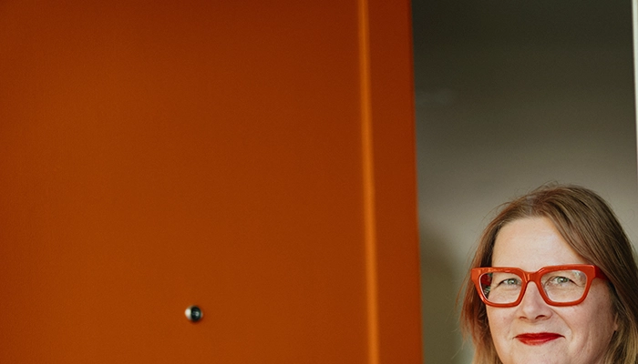 Woman with red glasses and red lips holding open orange door and smiling.