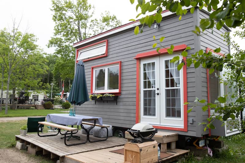 Small kit home red trim and double-doors. A small porch with a picnic table sits in front.