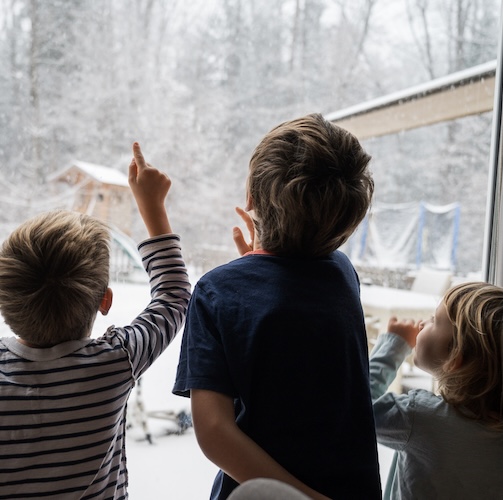 Three young children looking at snow out the window.