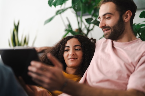 Couple looking at homes together on a tablet while snuggling on their couch.