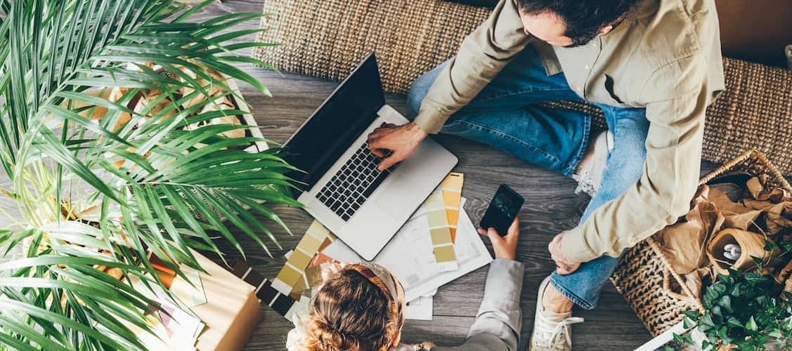 A couple sitting on the floor, engrossed in their work on a laptop, possibly related to the renovation of their new house.