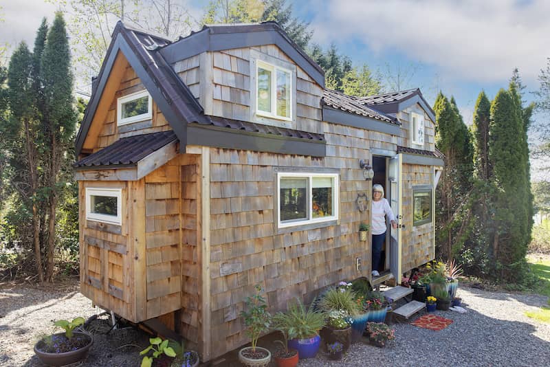 Friendly mature woman standing at front door of her charming little wooden tiny home.