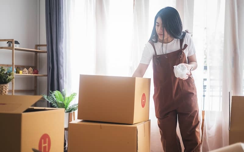 Woman in overalls packing moving boxes in living room.