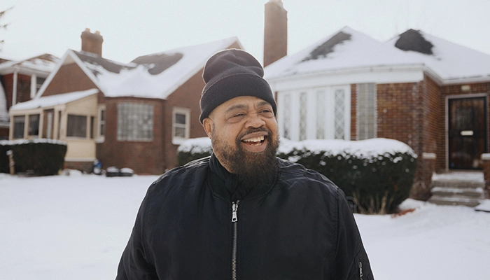 Man standing in the snow in front of brick house wearing black beanie and black zip up coat.