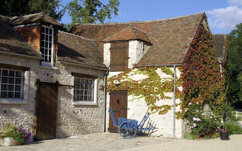 French cottage made of light stone, dark brown doors and large windows with climbing greenery on its front and side.