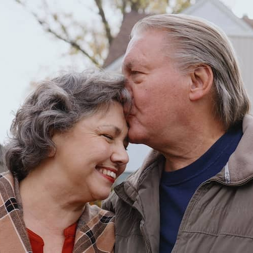 Man kissing forehead of smiling wife in front of home.