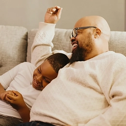 A man wearing glasses laughs while reclining on a couch as a young boy leans against him, smiling and laughing too. They appear relaxed and playful in a cozy living room setting.