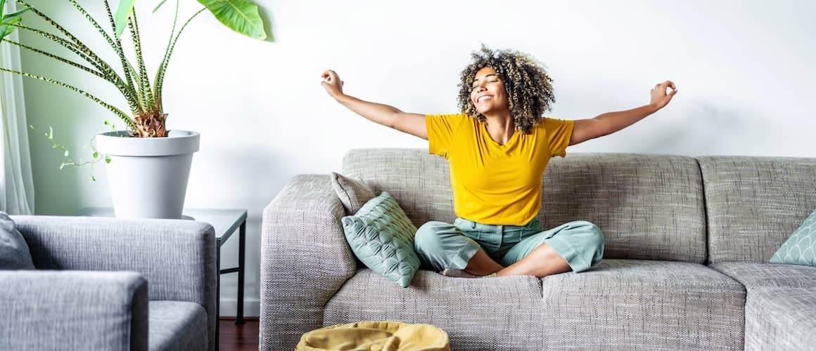 Woman smiling, siting in lotus position on couch in living room of new home, with arms outstretched in joy.