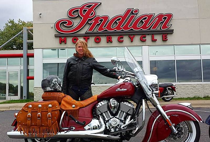 Woman standing behind her red motorcycle in front of a building that reads "Indian Motorcycle."