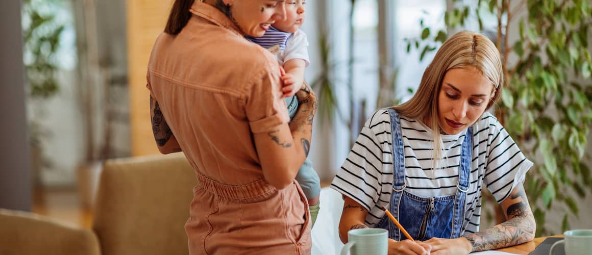 Young mothers budgeting together - one is standing up holding their baby and the other is writing down information at the kitchen table.