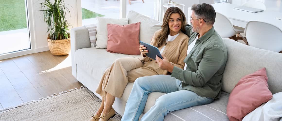 Middle aged couple sitting on couch, holding a tablet and looking at each other.