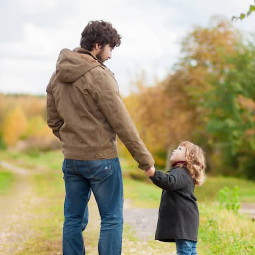 Smiling father in tie affectionately walking down tree-lined street holding hands with his young daughter.