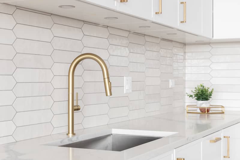 White picket tile backsplash behind gold faucet over inlaid sink in white countertops.