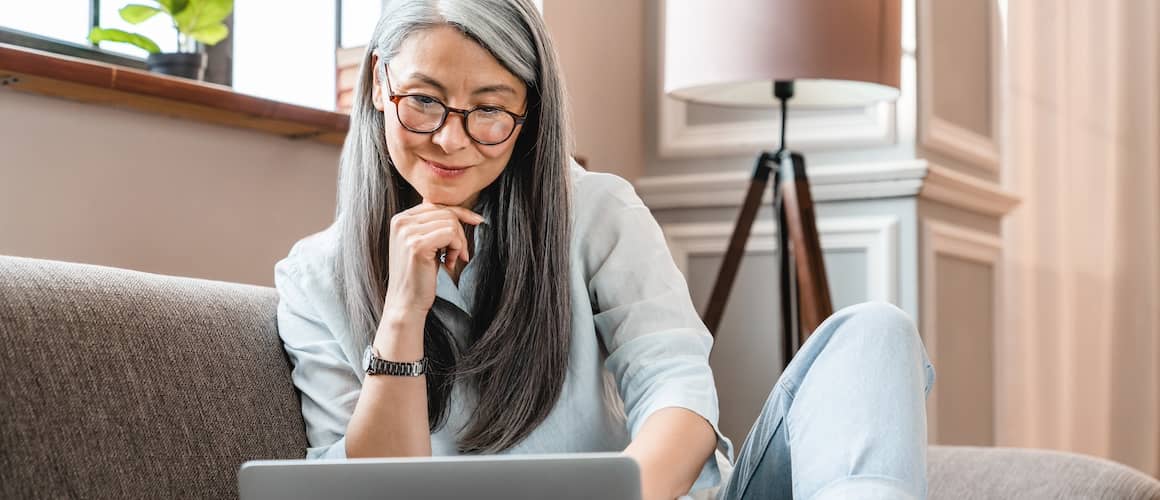 Older woman wearing glasses, sitting on grey couch and looking at open laptop.