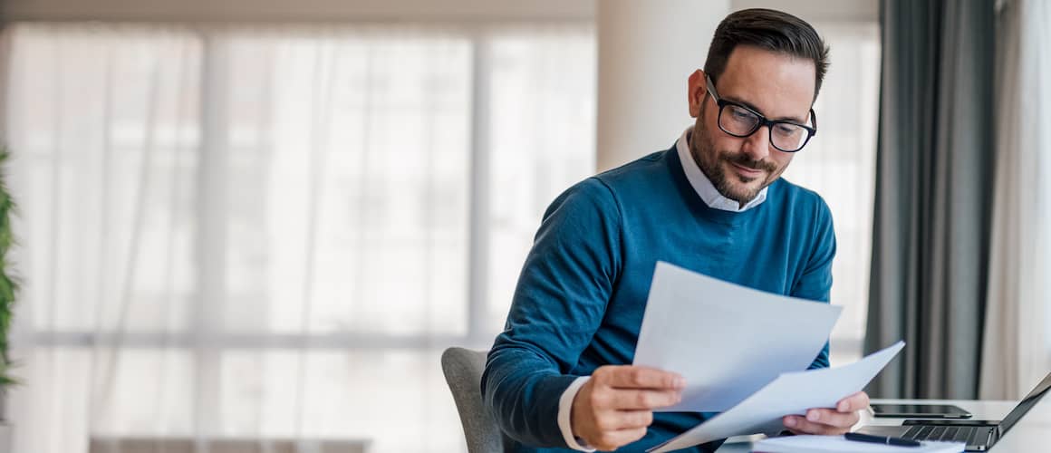 Man at home sitting at a desk and looking at papers in his hand.