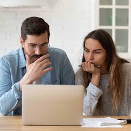 Couple sitting at the kitchen table and staring intently at a laptop. 
