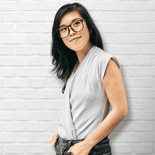 Woman wearing glasses smiling in front of a white brick wall.