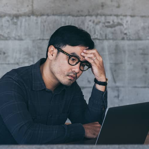 A young Asian man looking stressed while using a laptop, possibly in a home or work setting.