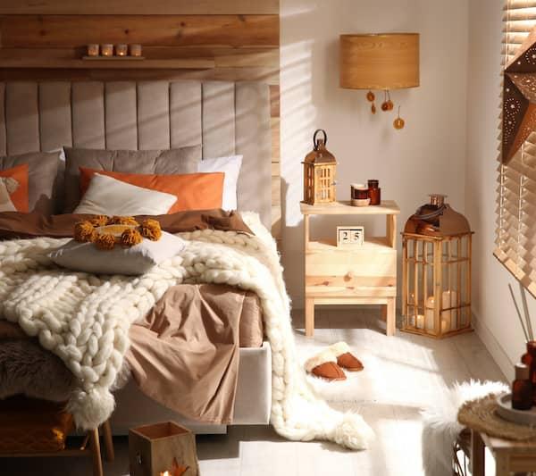 A sunlit cozy bedroom with a knitted blanket and comfortable looking pillows scattered about.