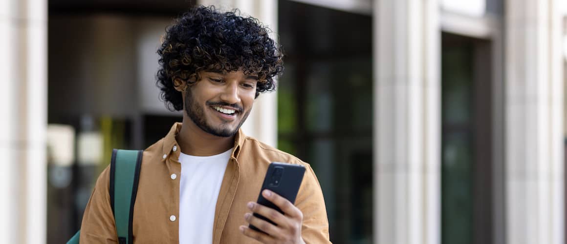 Man smiling at phone while walking through university campus with books.