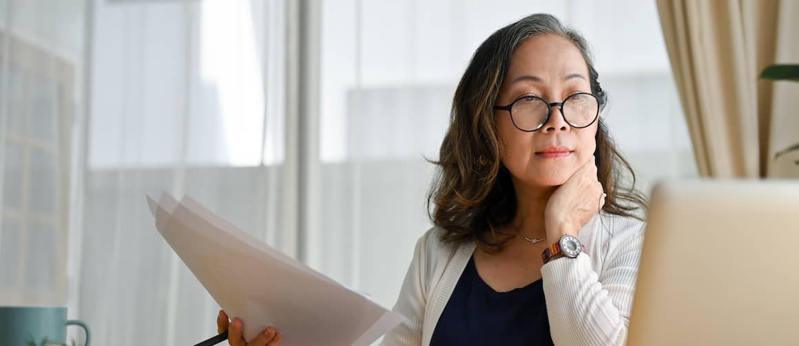 Middle aged woman wearing glasses, holding documents and looking at laptop.