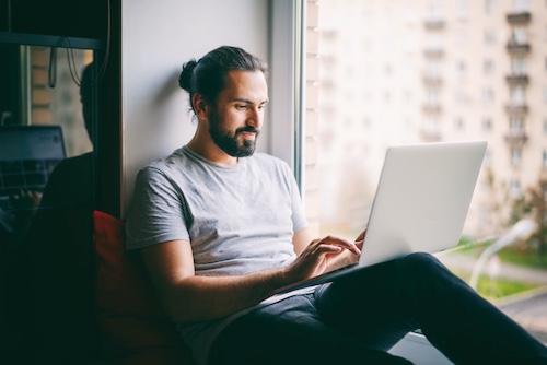 Young man sitting in widow seat of apartment on laptop.