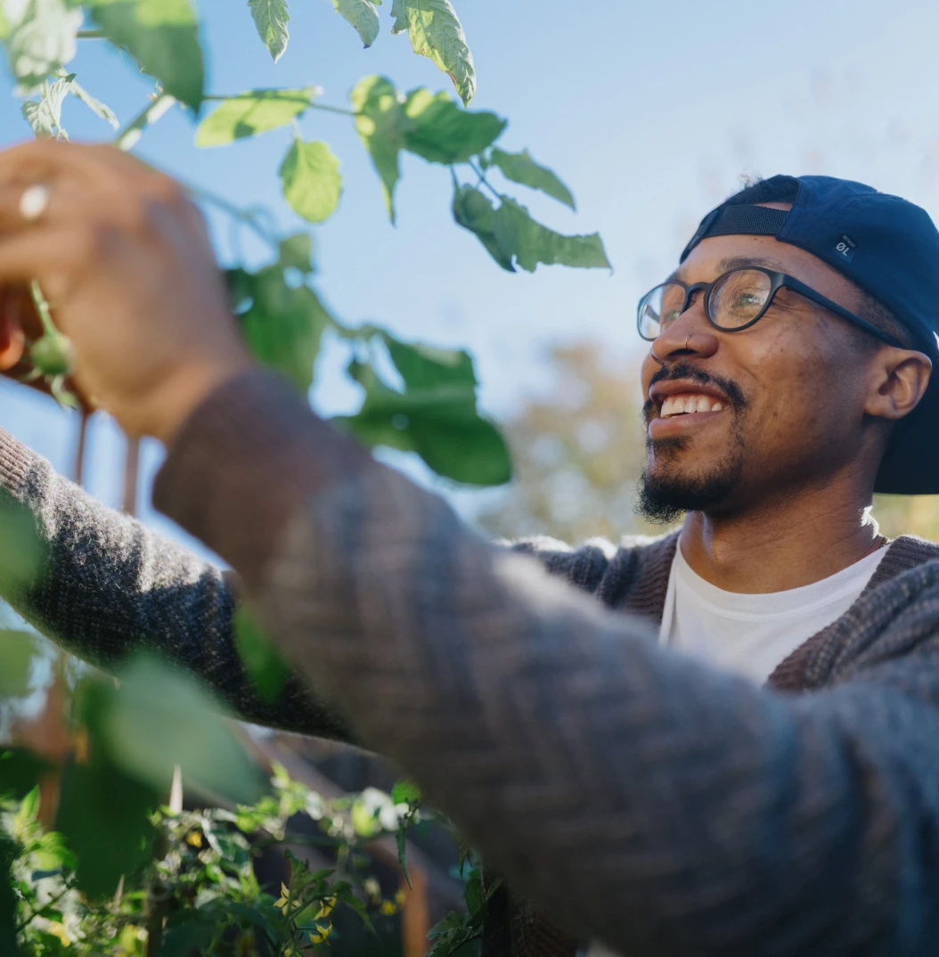 A man wearing glasses and a hat smiles while tending to plants in a garden on a sunny day.