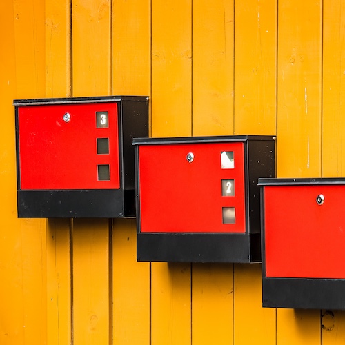Staggered orange artistically displayed mailboxes on yellow panelled exterior of home.