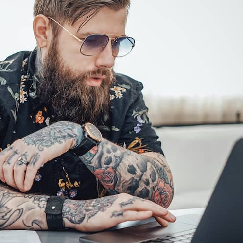 A stylish young man with tattoo sleeves working on a computer.