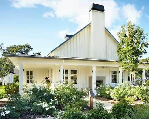White house with large wrap around covered porch and lush landscaping.